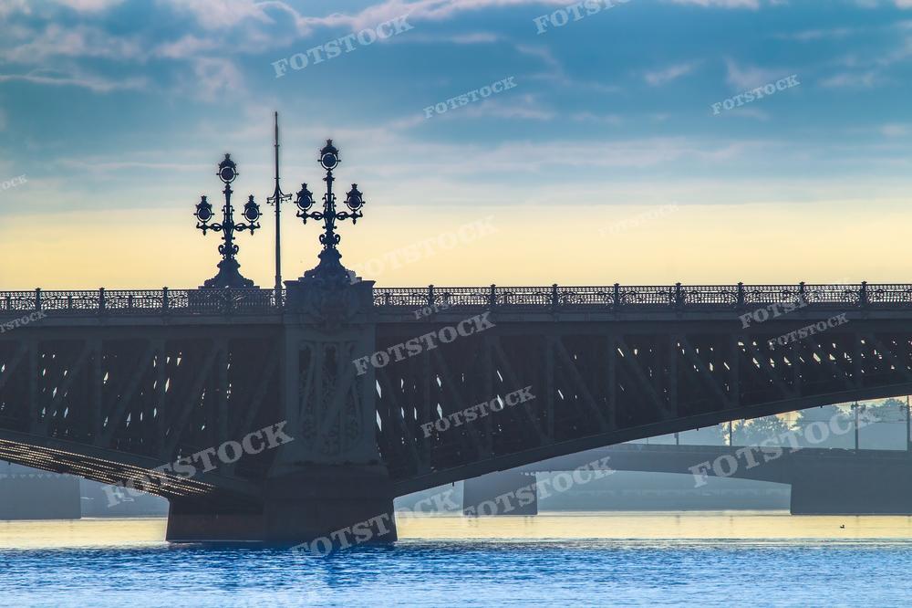 На фотографии изображен один из мостов Санкт-Петербурга. Троицкий Мост виден на фоне неба, освещённого утренним или вечерним светом, что придаёт изображению контрастность и драматизм. Фонари на мосту выполнены в характерном для города украшенном стиле. Это фото может использоваться для туристических буклетов, иллюстраций путеводителей по Санкт-Петербургу, в качестве обложки книги или альбома о городе, а также в статьях о российской архитектуре и инженерных сооружениях.